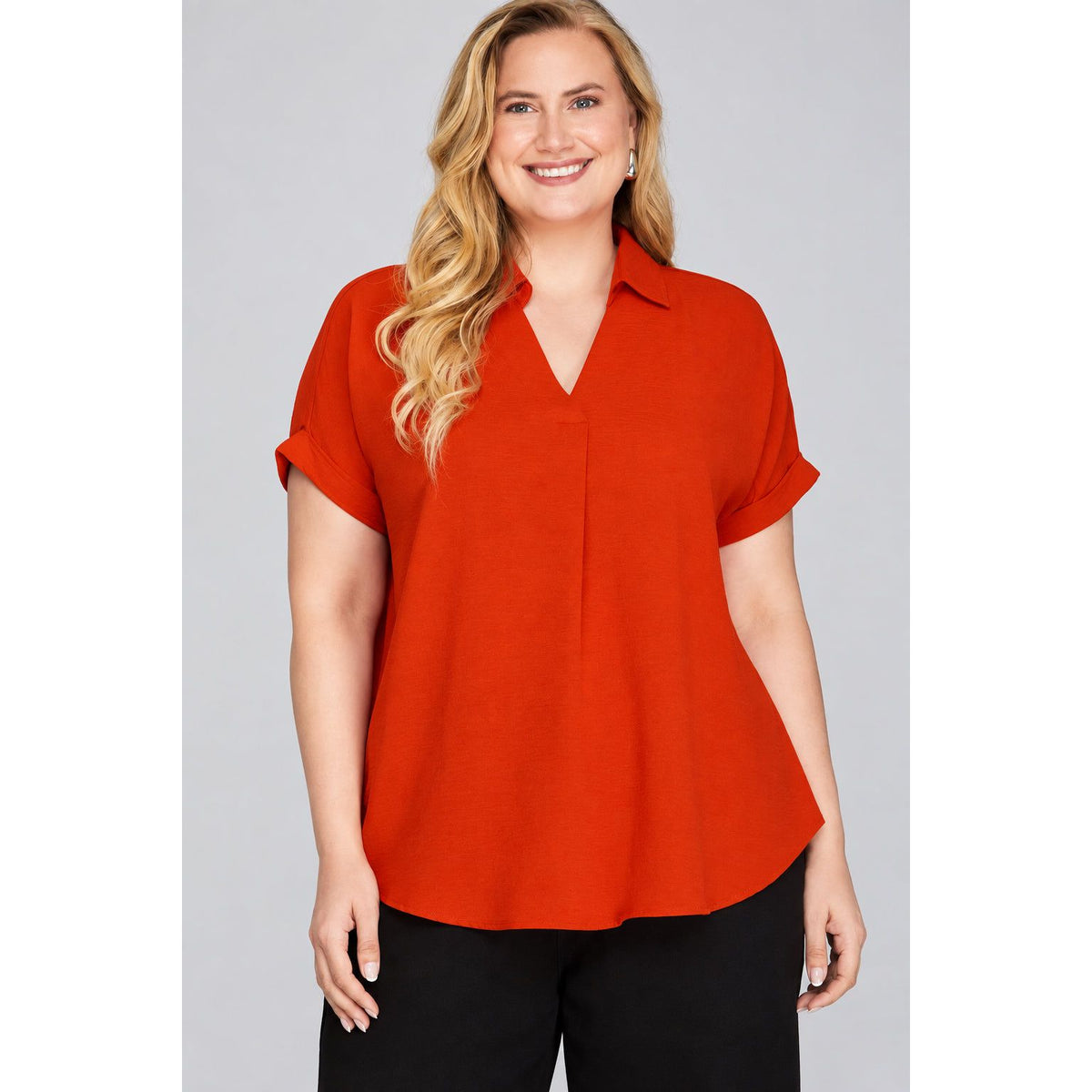 A woman modeling a short-sleeve collared blouse in a vibrant rust orange color, paired with black pants, against a solid light gray background.