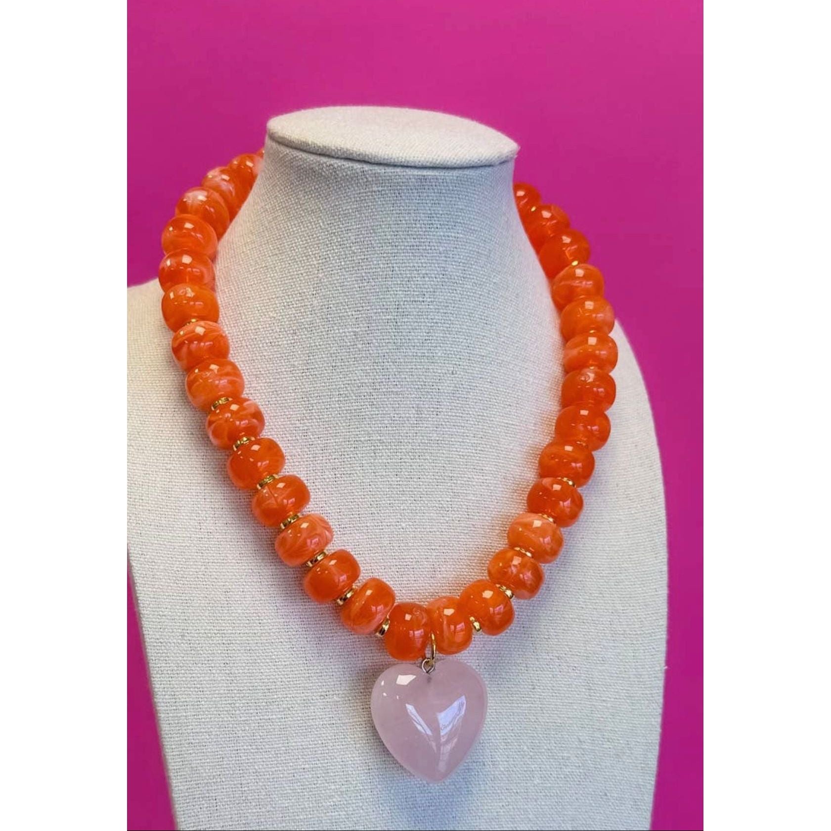 Necklace with orange beads and a pink heart pendant on a mannequin bust against a pink background