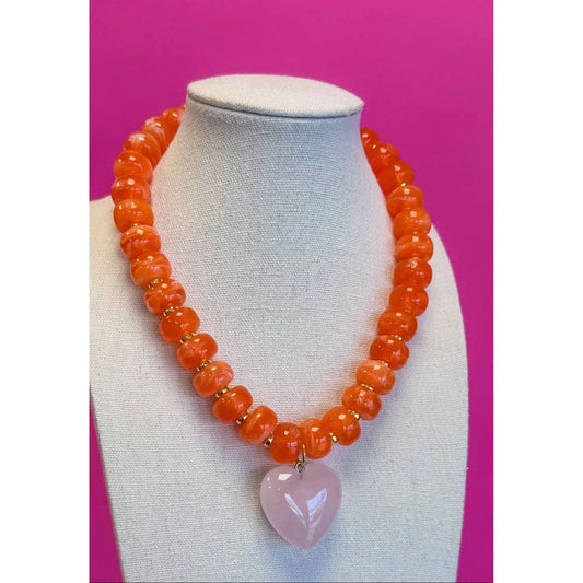 Necklace with orange beads and a pink heart pendant on a mannequin bust against a pink background