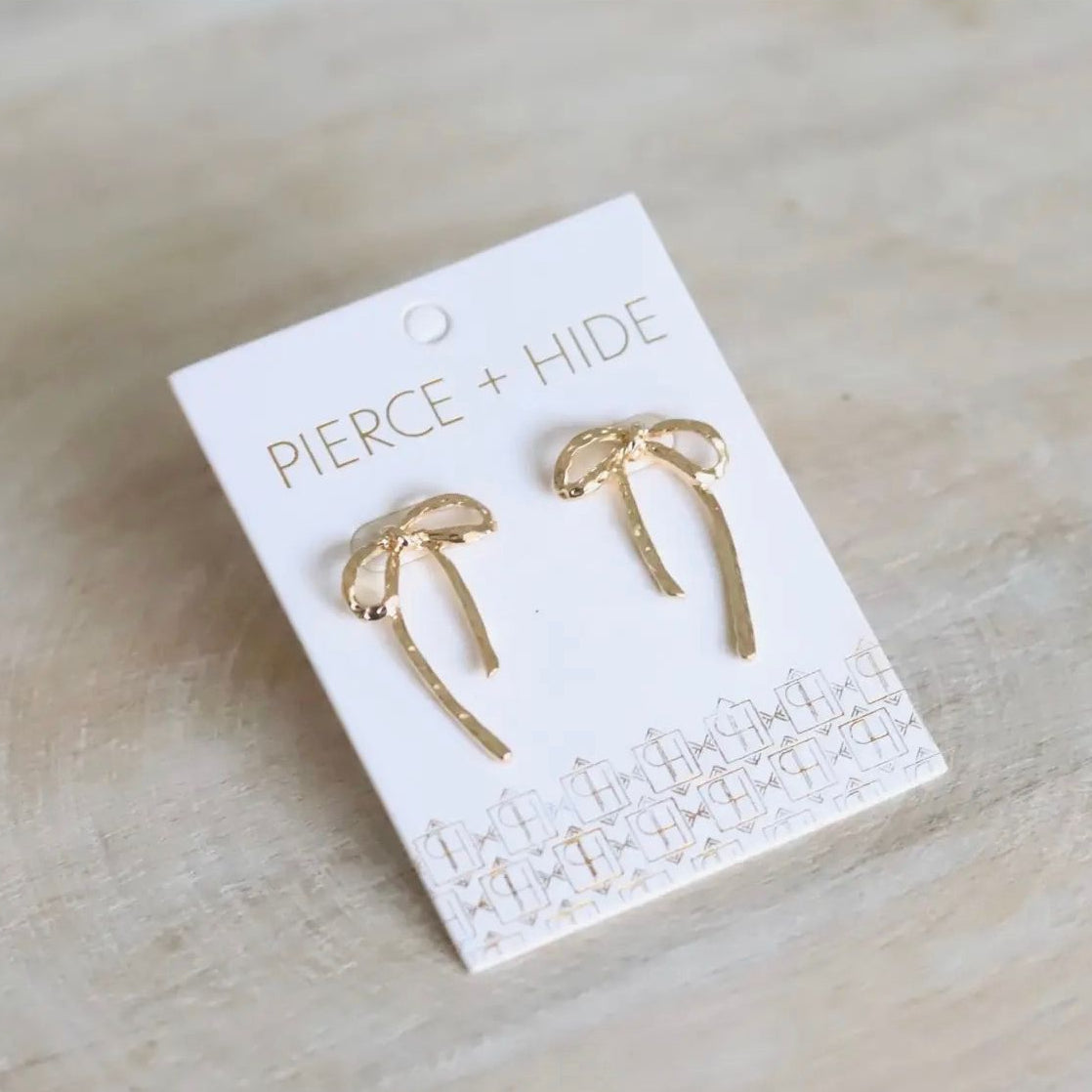 Gold bow-shaped earrings on a 'Pierce + Hide' branded card against a beige background