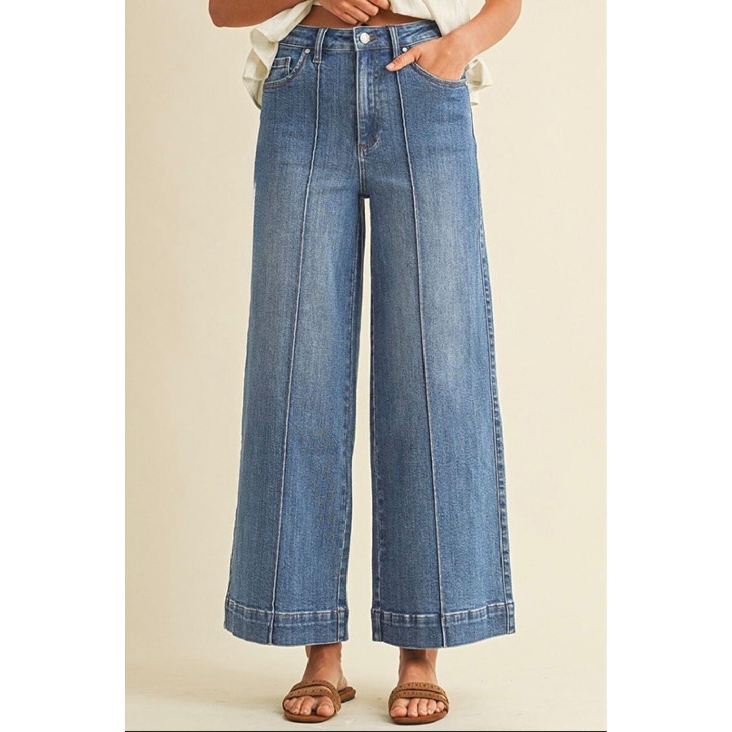 A person wearing high-waisted, wide-leg blue denim jeans with a straight-to-flared leg and rolled hems, paired with brown slide sandals against a solid beige background.