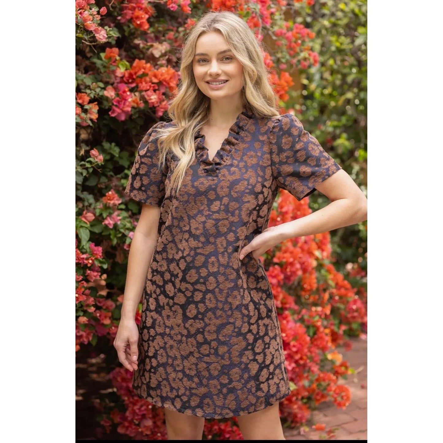 A woman wearing a short-sleeve mini dress with a brown and navy blue abstract pattern, standing outdoors in front of a background of red flowers and green foliage.