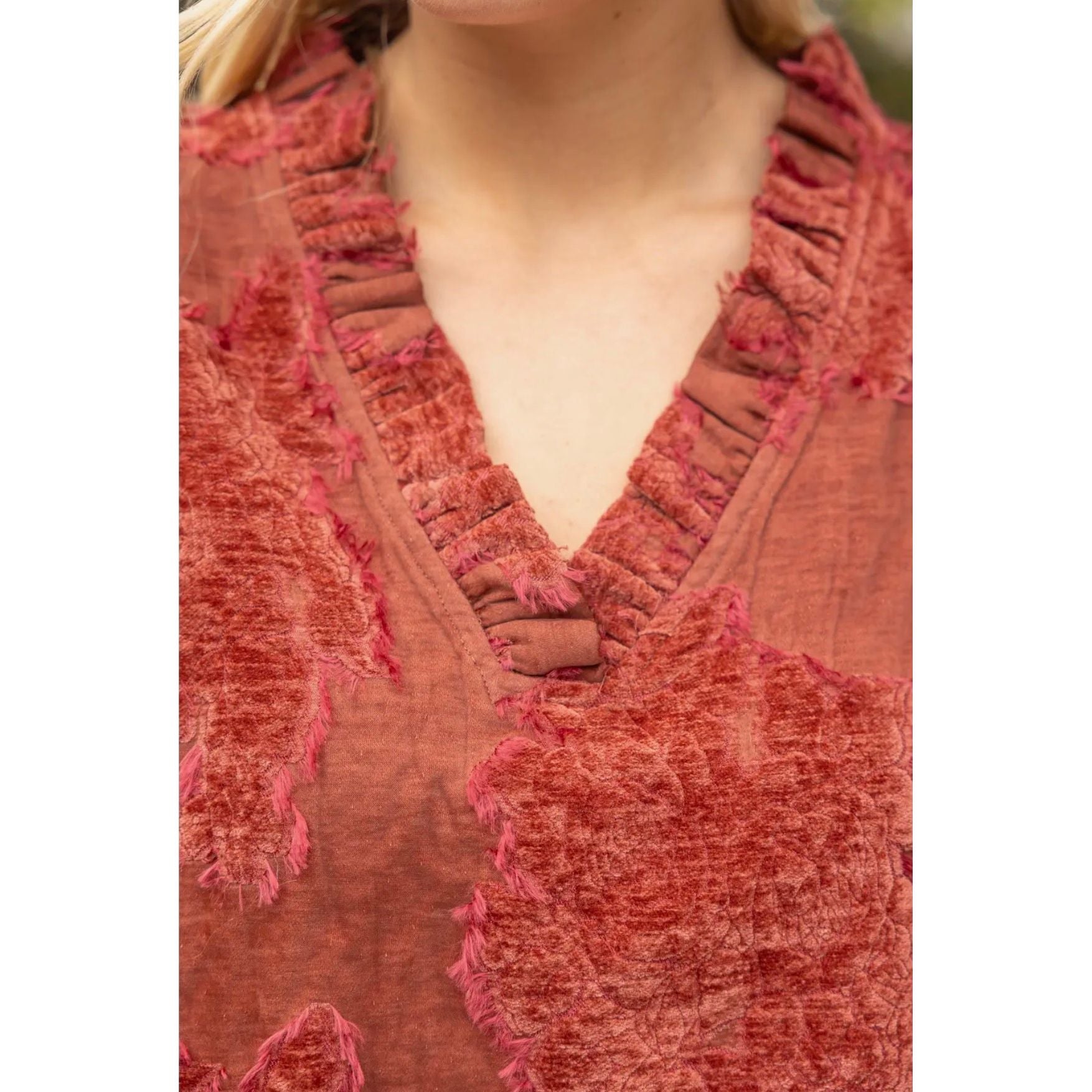 Close-up of a textured terracotta red top with a ruffled V-neckline and raw fringe details, worn by a model.