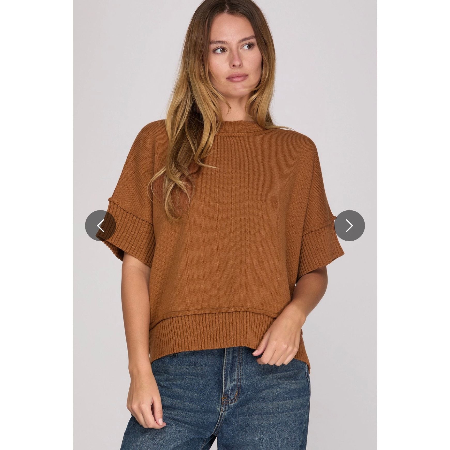 A woman wearing a brown short-sleeve knit sweater with ribbed cuffs and hem, paired with blue jeans, against a light gray studio background.