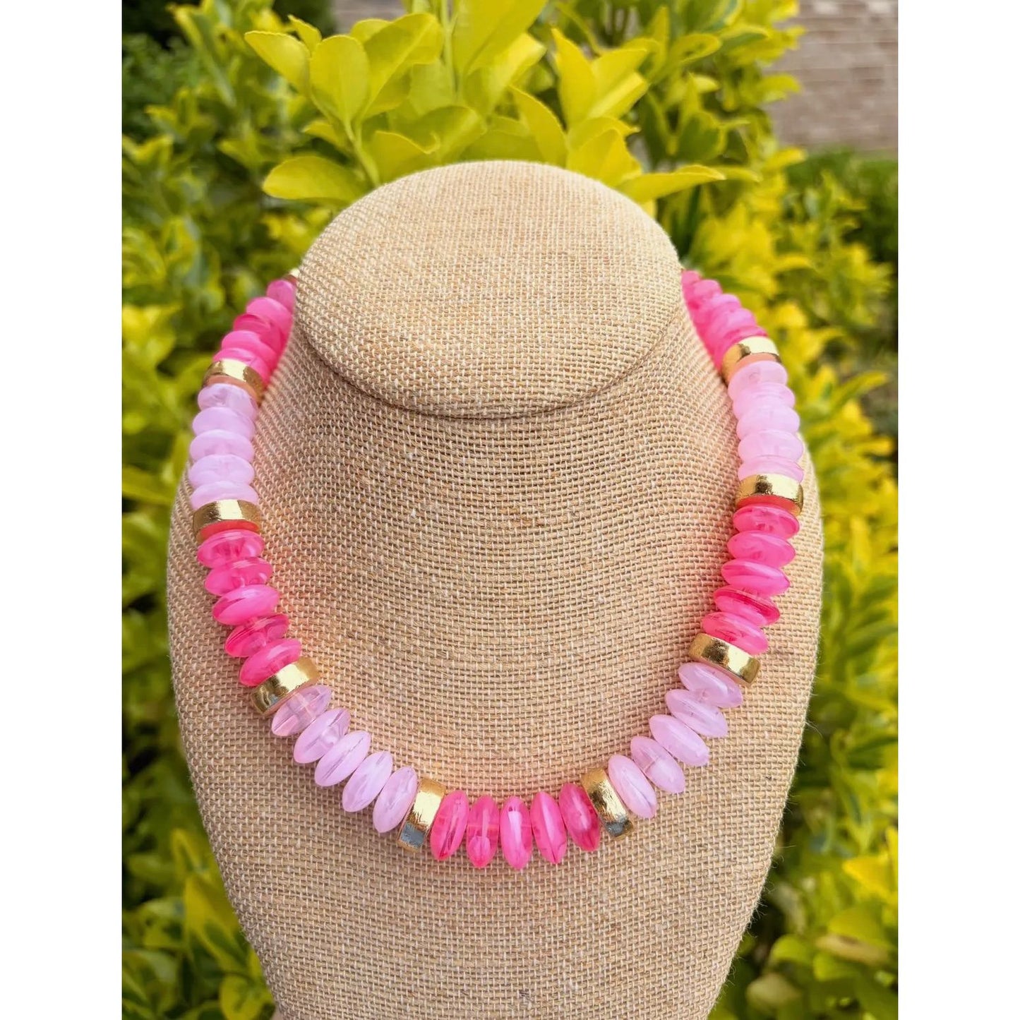 Necklace with pink and gold beads on a beige bust against a green foliage background