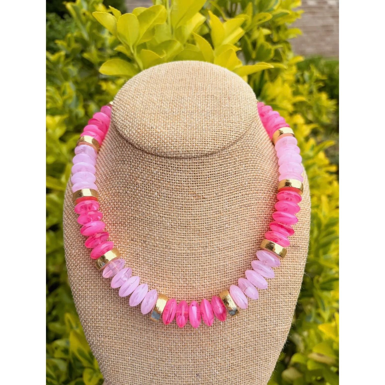 Necklace with pink and gold beads on a beige bust against a green foliage background