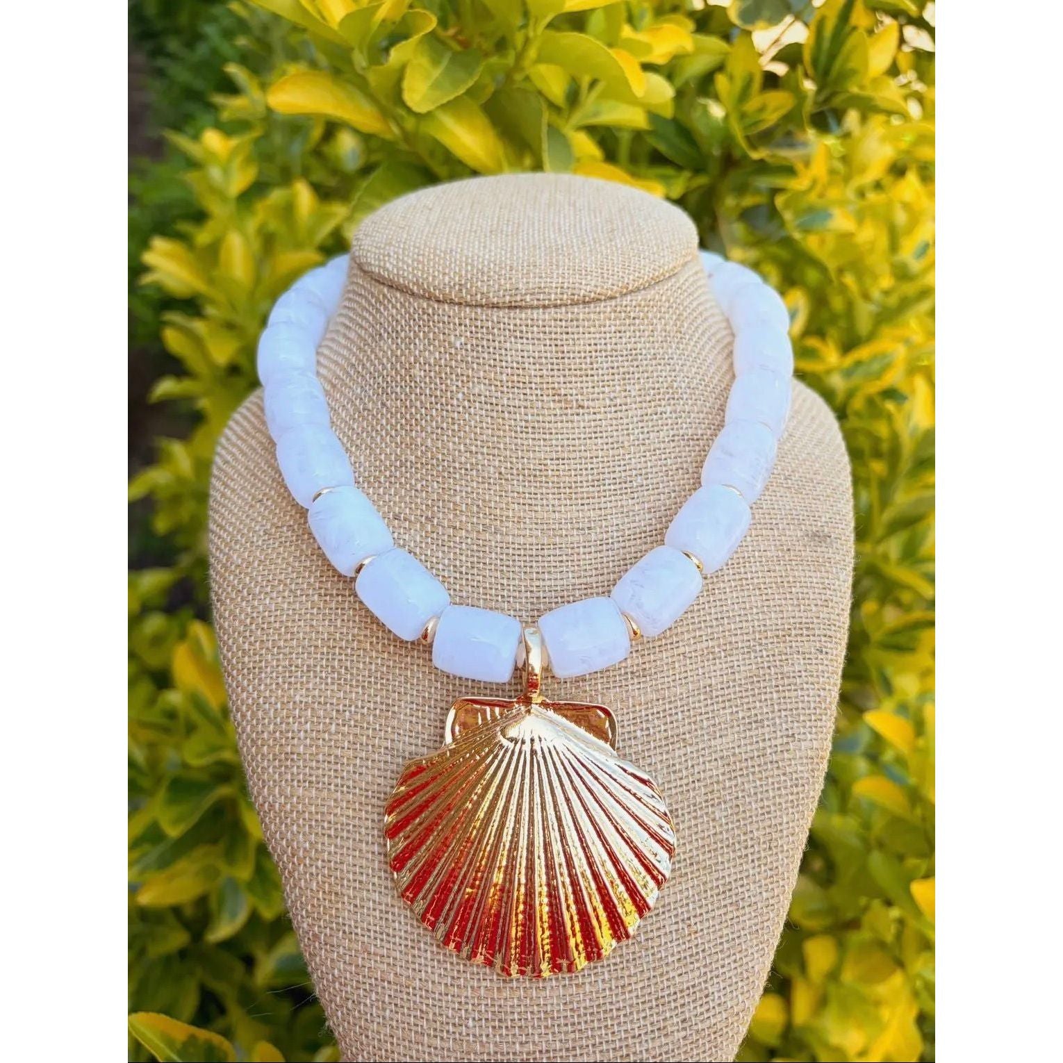 Necklace with light blue beads and a shell pendant on a mannequin bust against a green leafy background.
