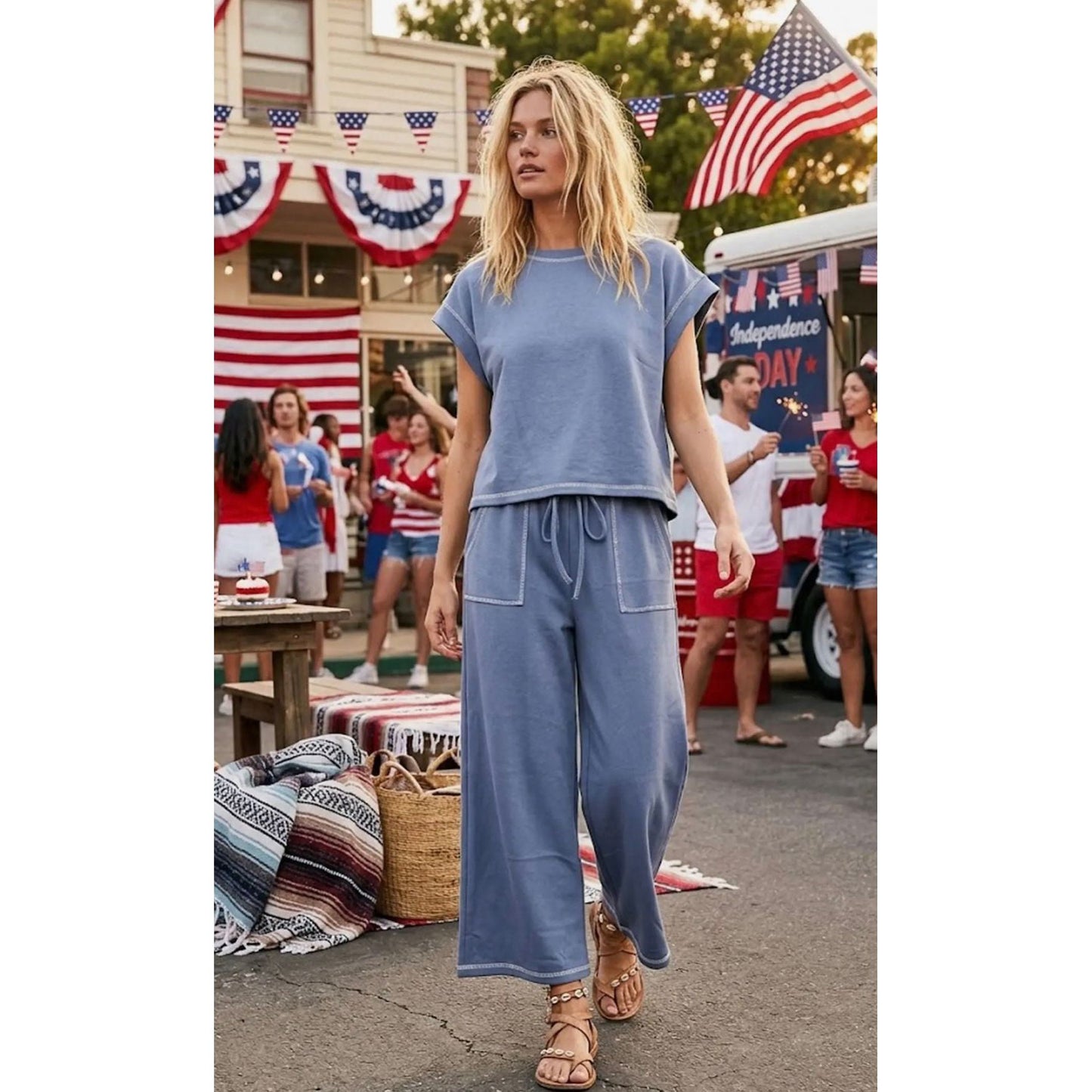 A woman stands outdoors at a patriotic parade or festival wearing a light blue short-sleeve top and matching wide-leg drawstring pants. Flags, banners, and other people are visible in the background.