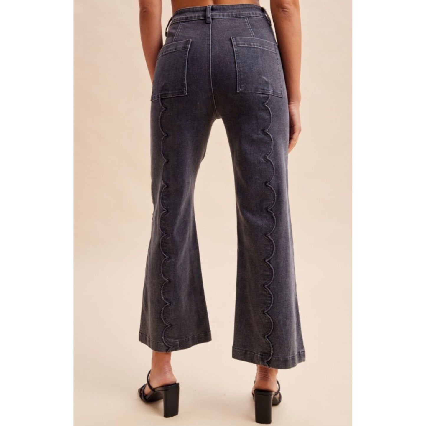 Back view of a person wearing dark gray wide-leg jeans with scalloped seam detailing down the outer sides, paired with black block-heel mules, against a plain beige studio background.