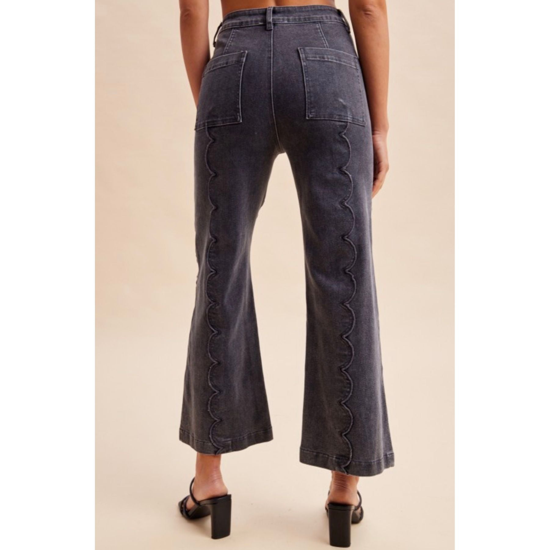 Back view of a person wearing dark gray wide-leg jeans with scalloped seam detailing down the outer sides, paired with black block-heel mules, against a plain beige studio background.