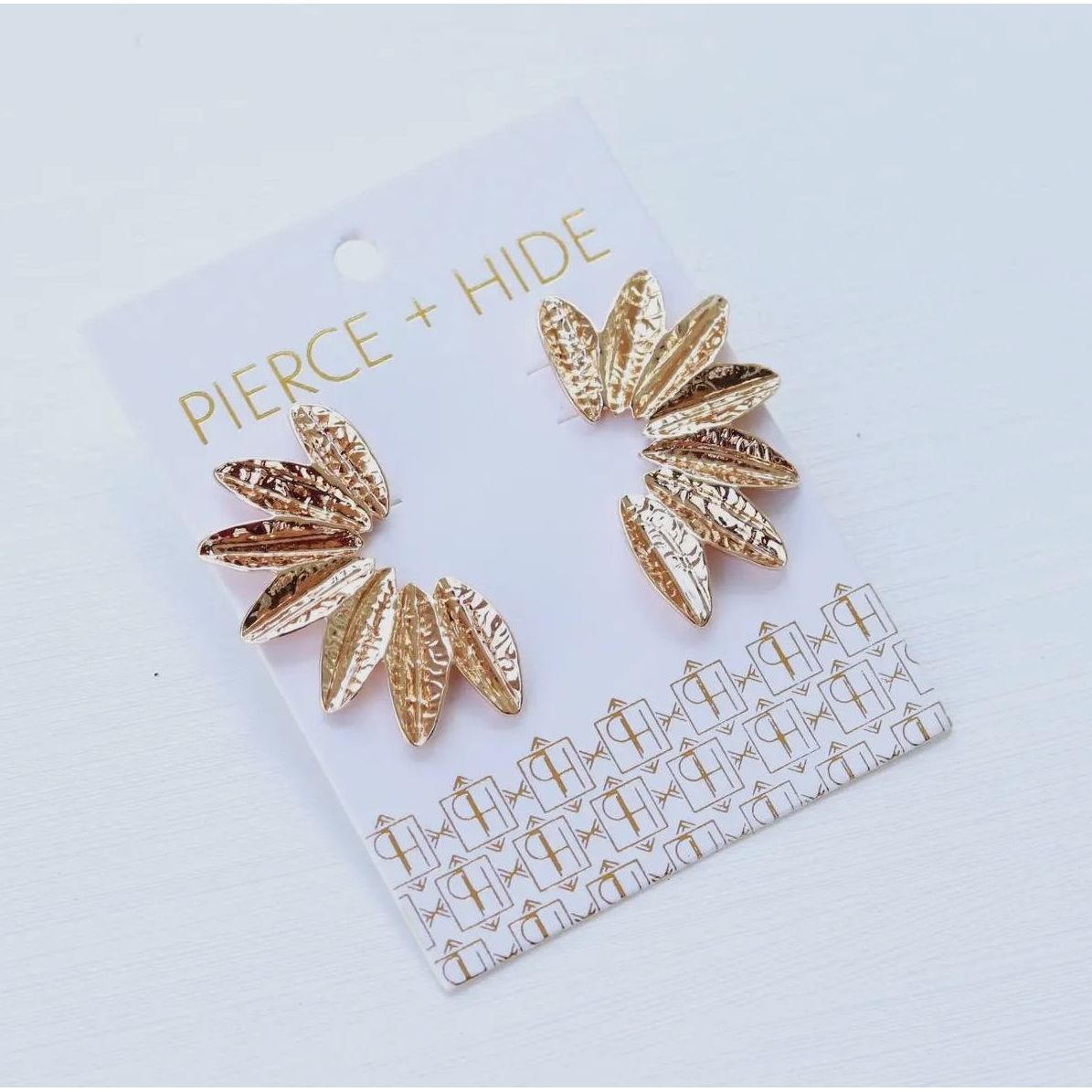 Gold leaf-shaped earrings on a 'Pierce + Hide' branded card against a light blue background
