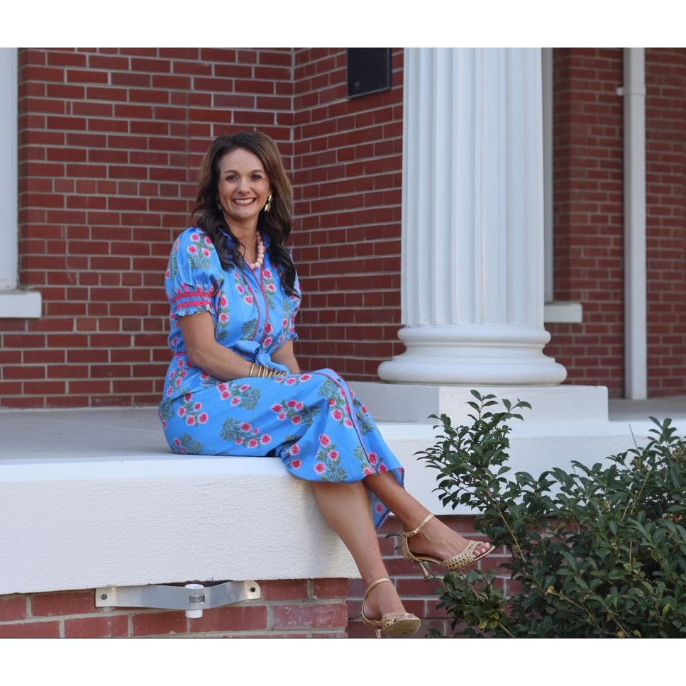 Woman in a blue floral dress sitting on a white chair with a brick wall and column in the background.