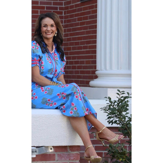Woman in a blue floral dress sitting on a white chair with a brick wall and column in the background.