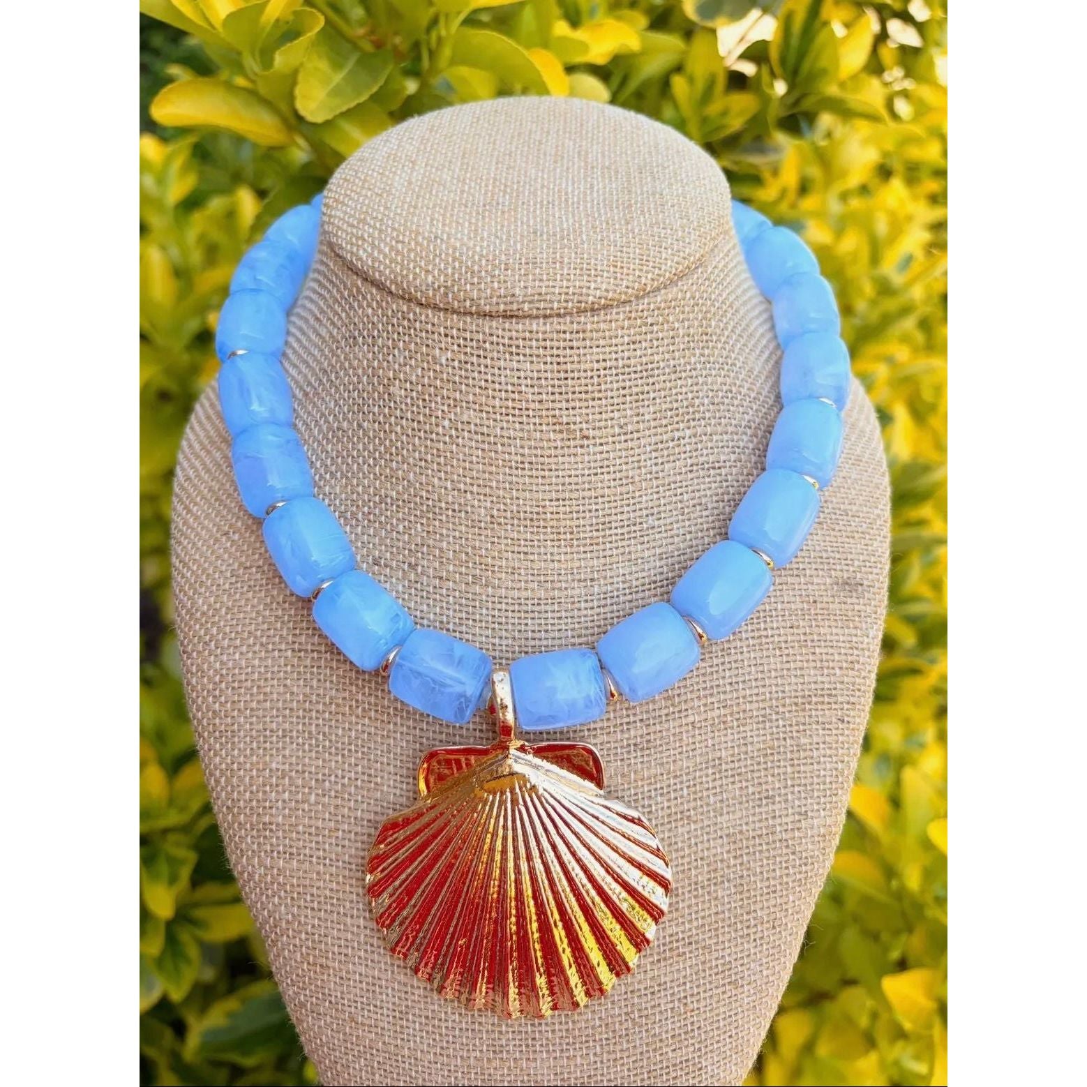 Necklace with blue beads and a gold shell pendant on a mannequin bust against a green leafy background.