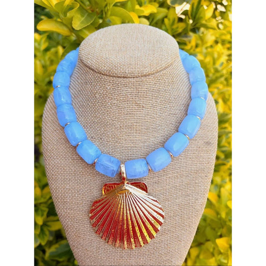 Necklace with blue beads and a gold shell pendant on a mannequin bust against a green leafy background.