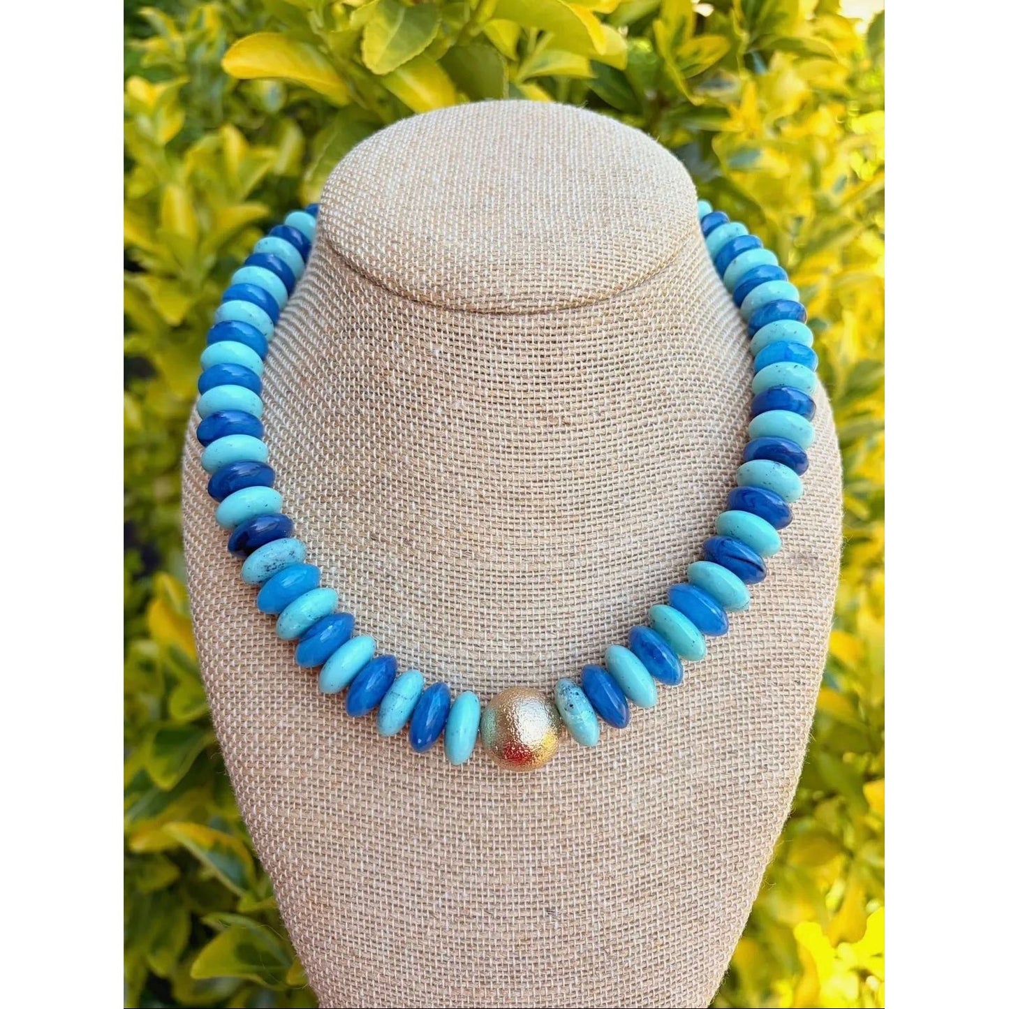 Necklace with blue and turquoise beads on a mannequin bust against a green leafy background