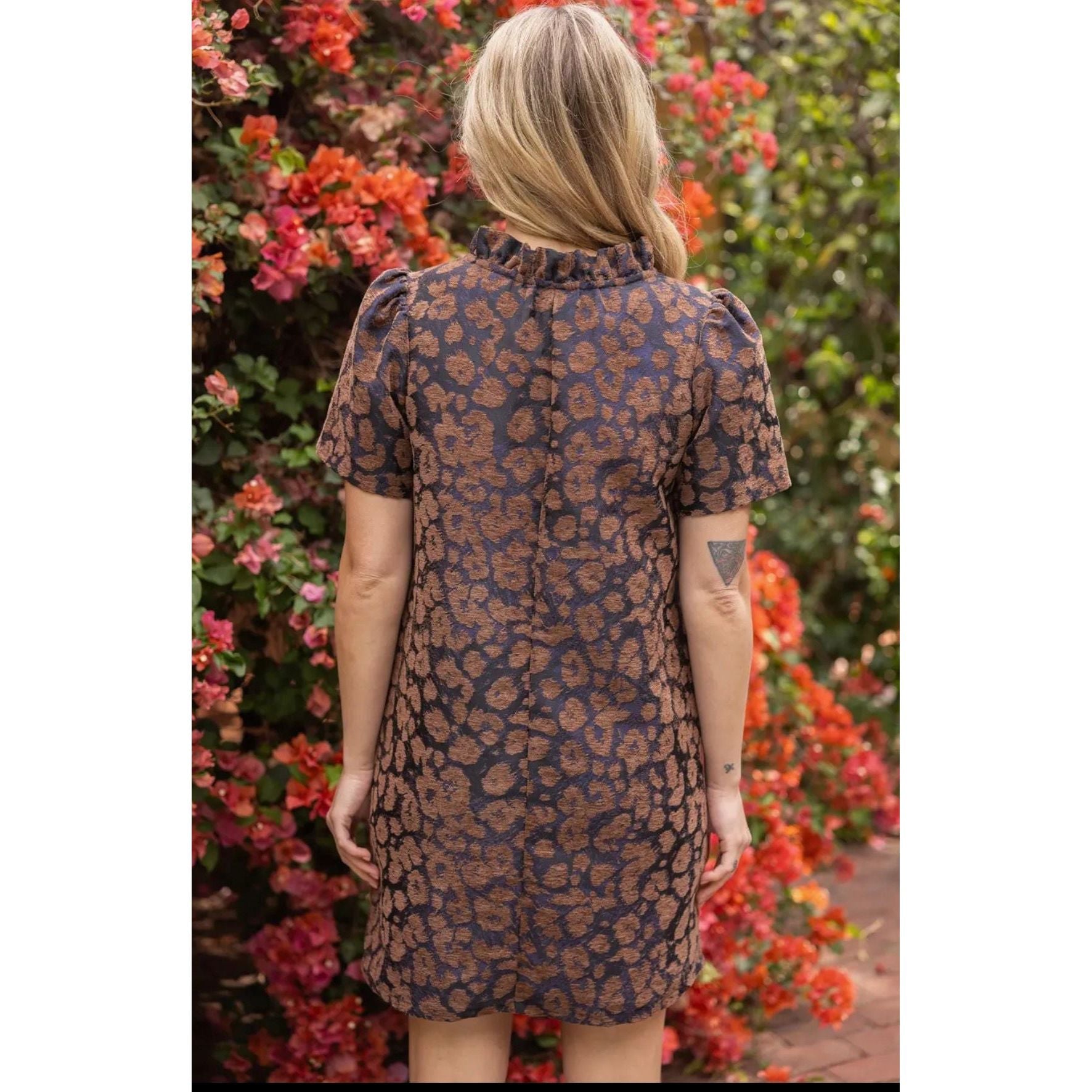 A woman viewed from behind wearing a short-sleeve dress with a brown and navy leopard print, standing in front of a background of red flowers and green foliage.