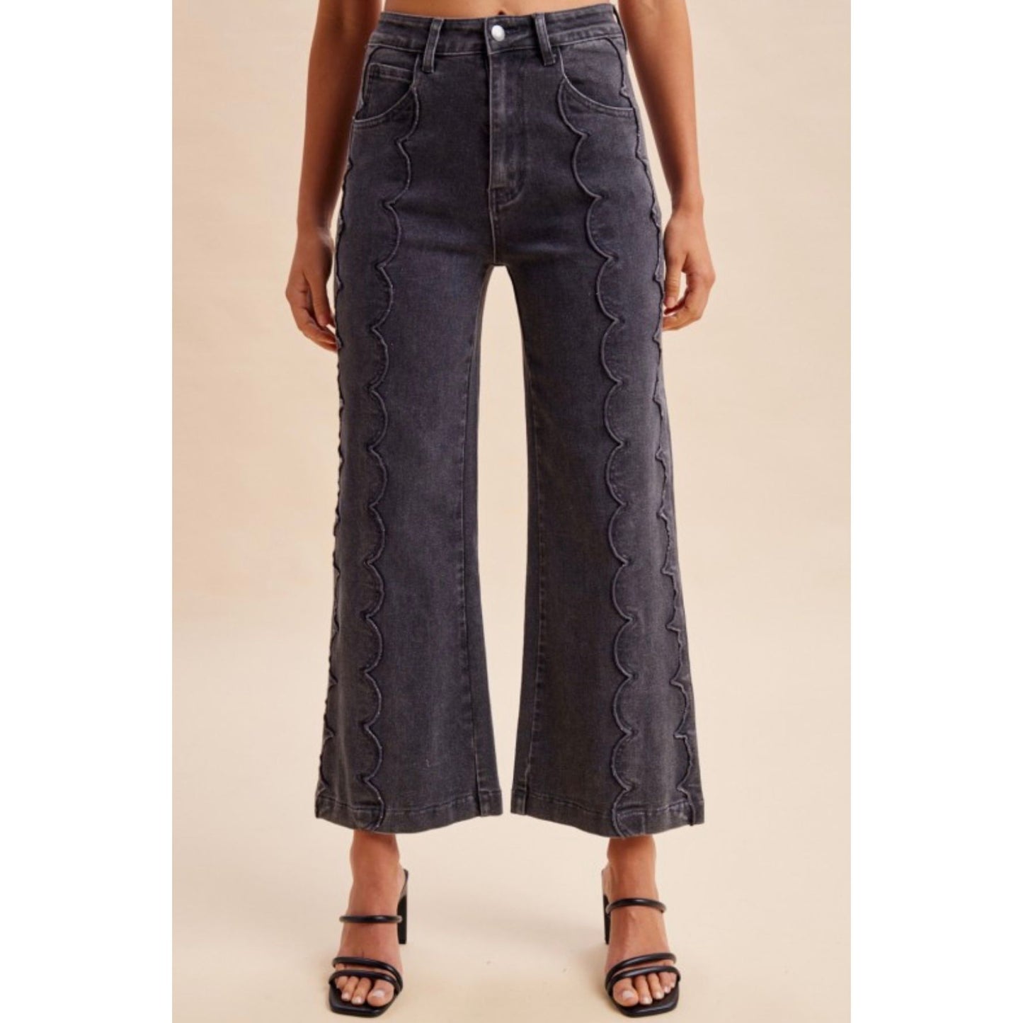 A model wearing dark gray wide-leg cropped jeans with vertical scalloped stitching down the sides, paired with black strappy high-heeled sandals against a solid beige background.