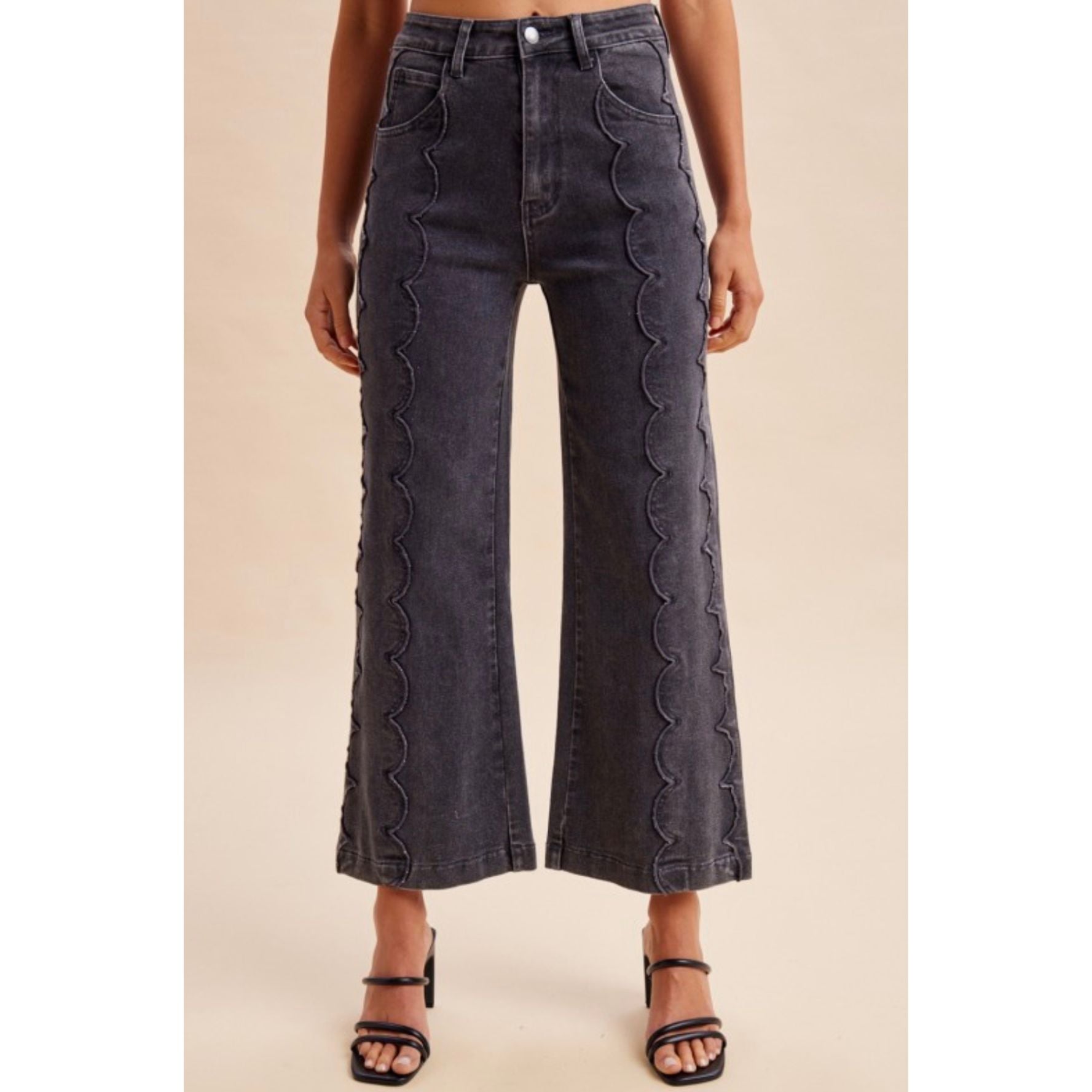 A model wearing dark gray wide-leg cropped jeans with vertical scalloped stitching down the sides, paired with black strappy high-heeled sandals against a solid beige background.