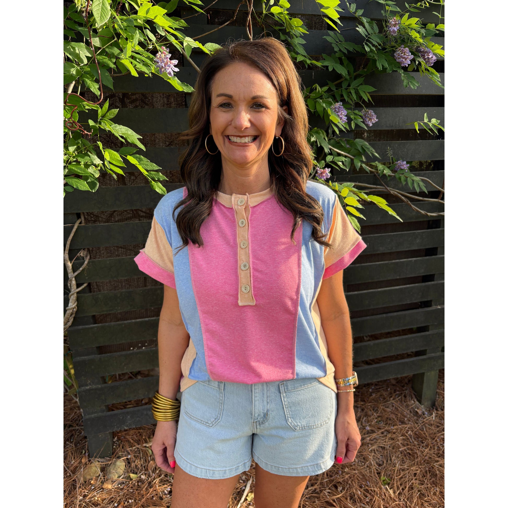 Woman wearing a colorful shirt and shorts standing in front of a wooden wall with greenery.