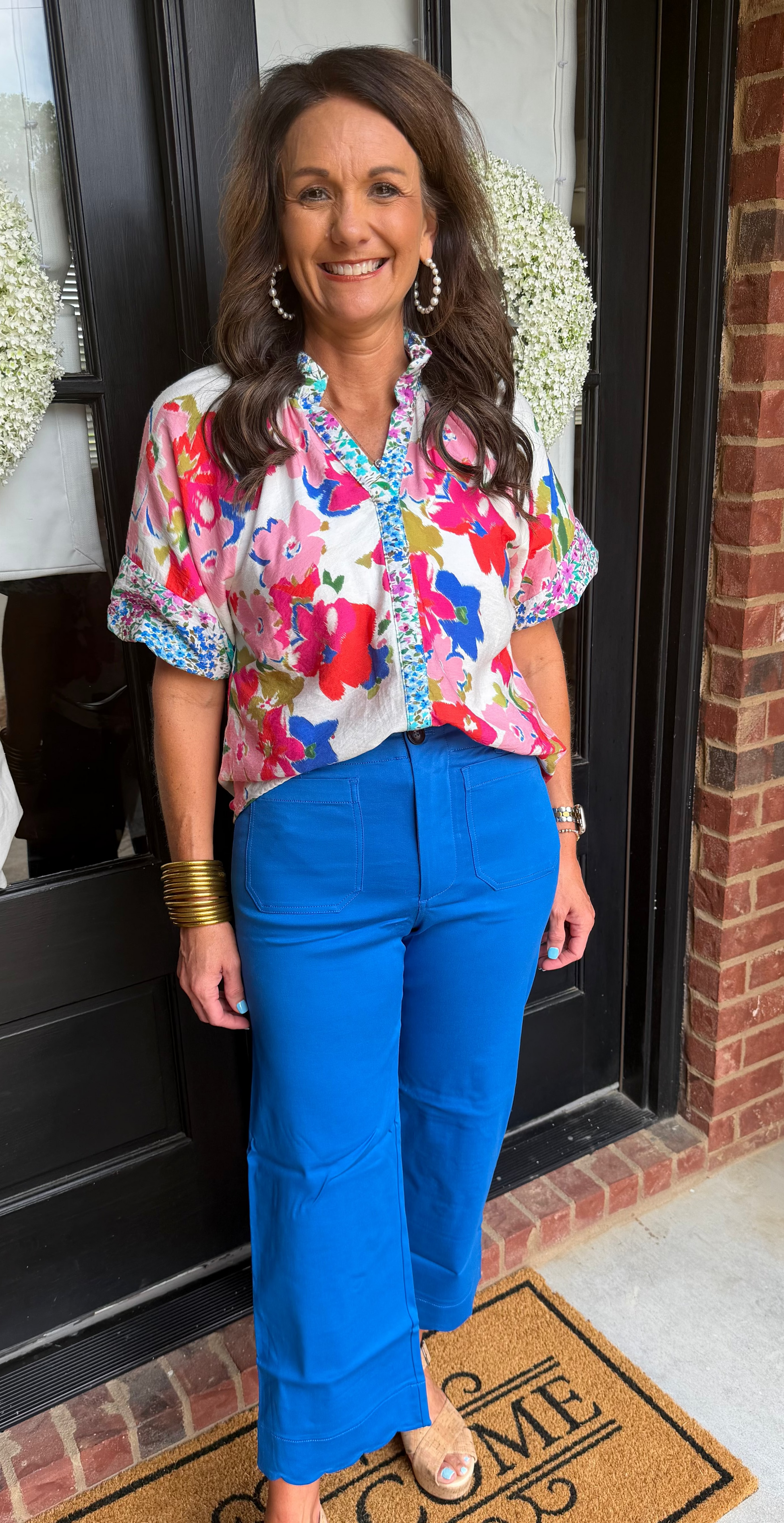 Woman standing in front of a door with a wreath, wearing a colorful blouse and blue pants.