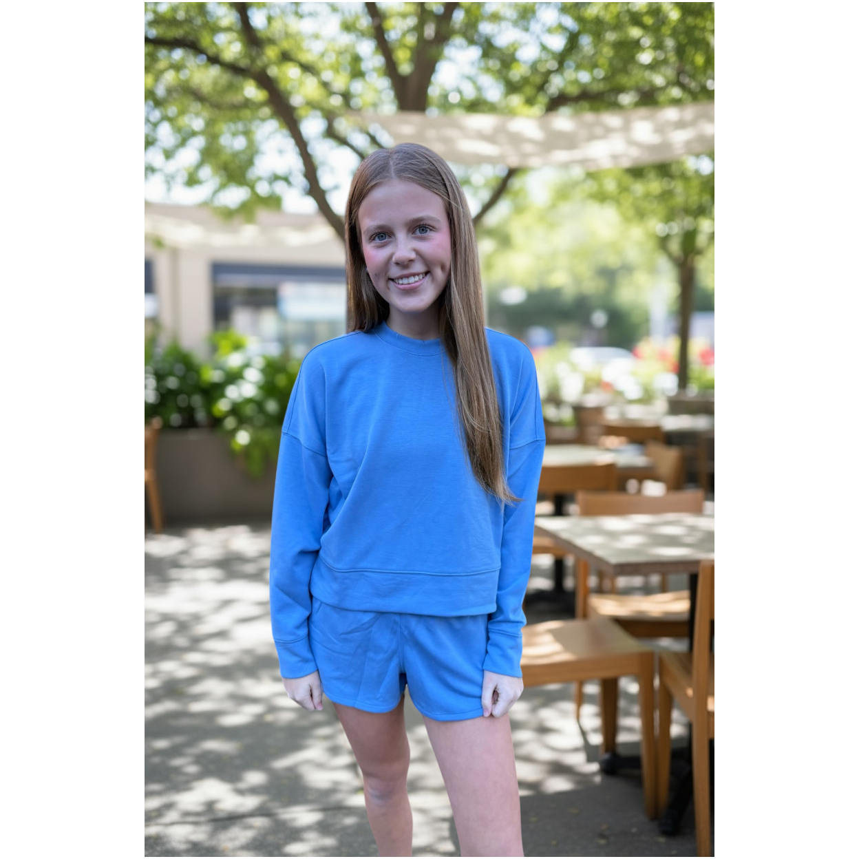 Young girl in a blue outfit standing outdoors with trees and tables in the background