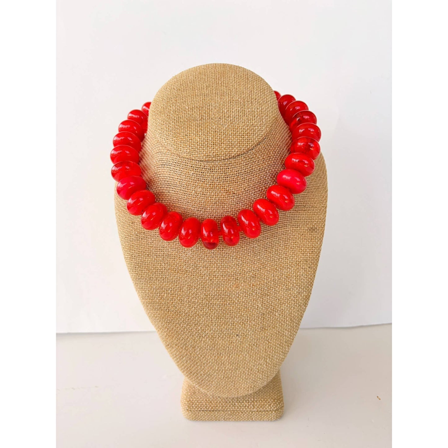 A chunky red beaded necklace displayed on a tan burlap jewelry bust against a plain white background.