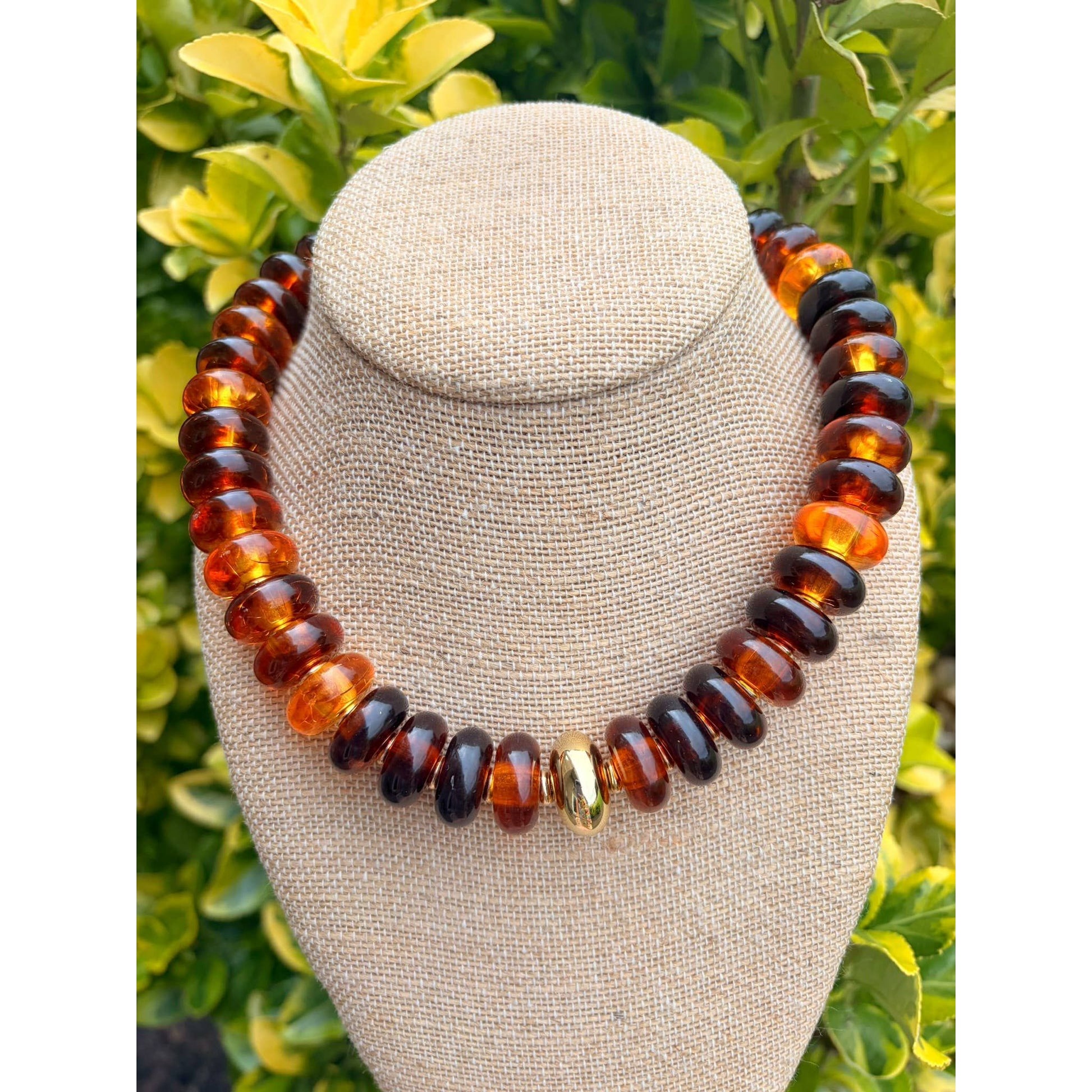 A chunky beaded necklace made of alternating amber-brown and dark black beads, with a single small gold accent bead in the center, displayed on a beige fabric jewelry bust against a background of green foliage.