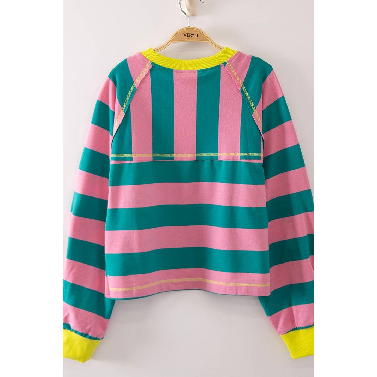 A pink and teal striped long-sleeve top hanging on a wooden hanger against a plain white wall. The shirt features bright neon yellow accents on the collar, sleeve seams, cuffs, and hem.