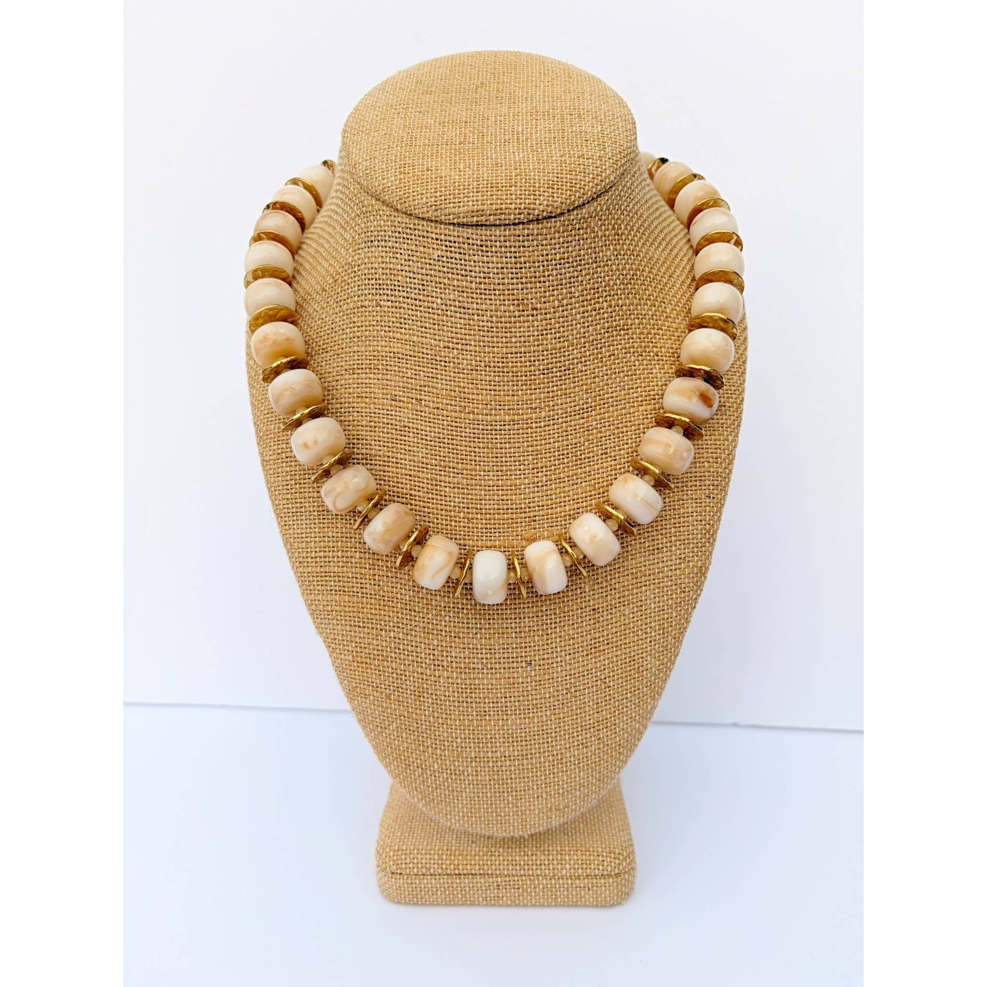 A necklace made of round cream-colored beads separated by small gold spacer rings, displayed on a tan fabric jewelry bust against a white background.