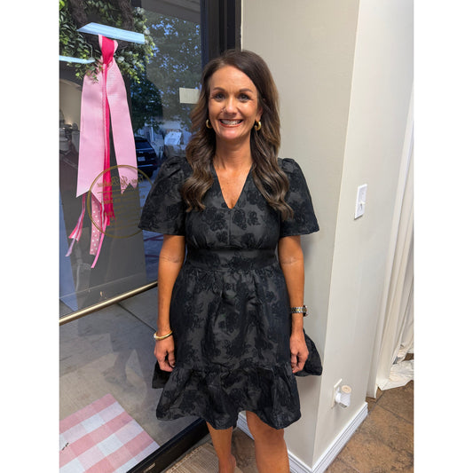 A woman wearing an Annie Ks Boutique black jacquard V-neck mini dress with puff sleeves and a cinched elastic waist.
