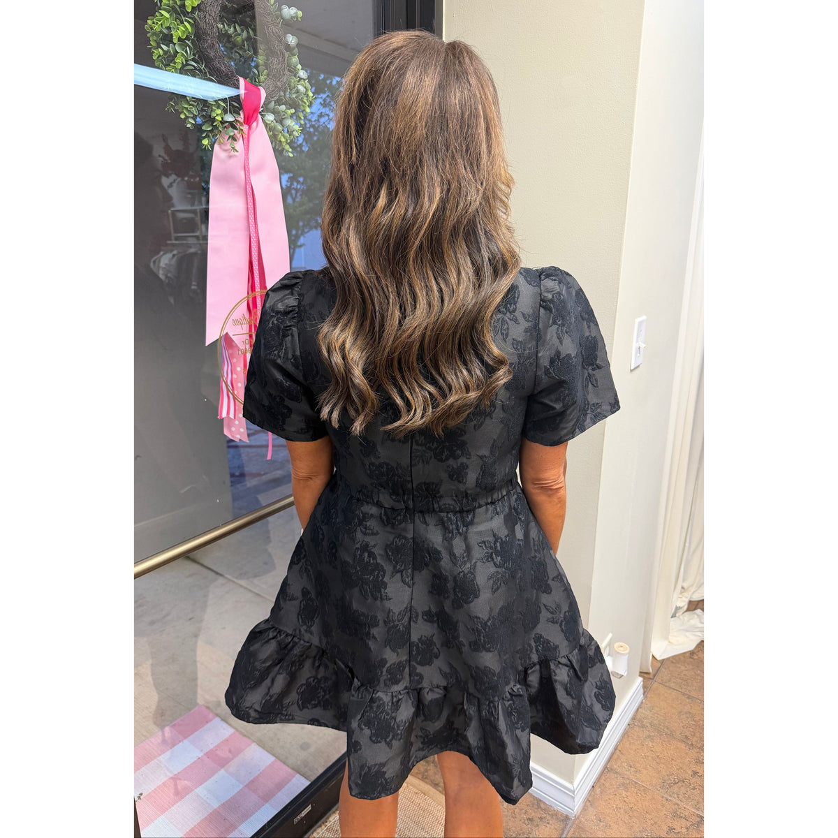 Back view of a woman wearing an Annie Ks Boutique black jacquard mini dress with short sleeves and a ruffled hem.