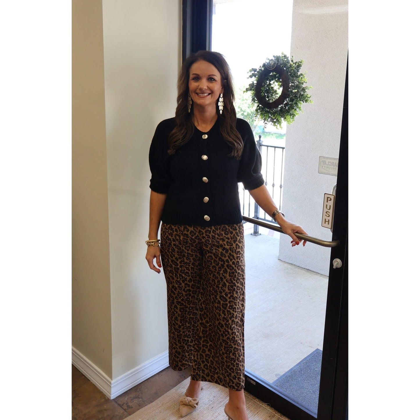 Annie Ks Boutique black puff sleeve sweater cardigan with gold buttons worn with leopard print pants by a doorway