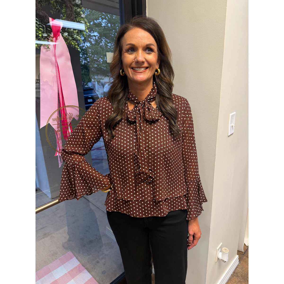 Annie Ks Boutique brown polka-dot top with bell sleeves and a neck bow tie worn by a woman in a store setting.