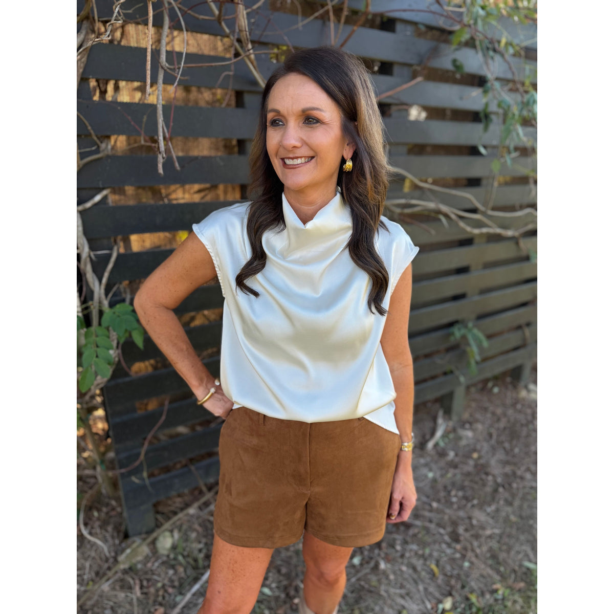 Annie Ks Boutique camel brown faux suede shorts paired with a white satin cowl neck top on a smiling woman