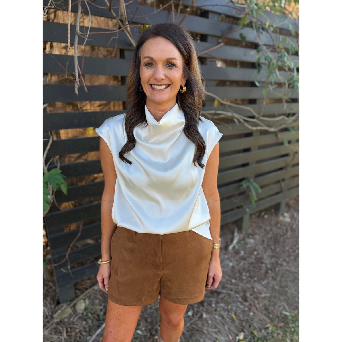 Annie Ks Boutique camel brown faux suede shorts paired with a white cowl neck satin top on a smiling woman