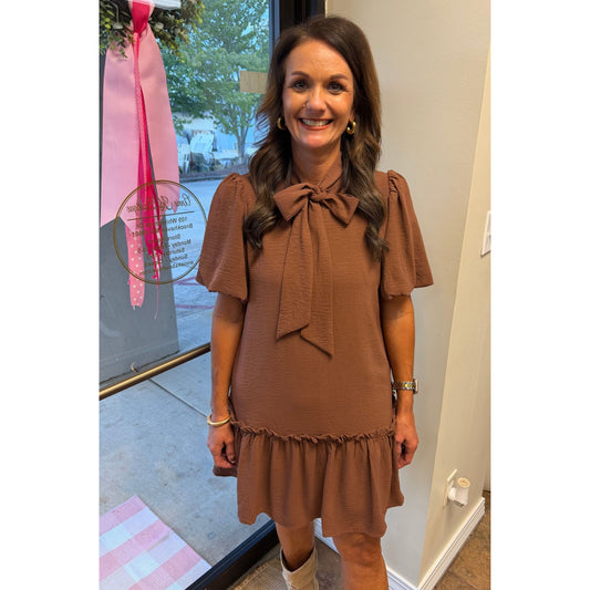 Woman wearing an Annie Ks Boutique caramel woven mini dress with a bow-neck detail and ruffled hem in a shop setting.