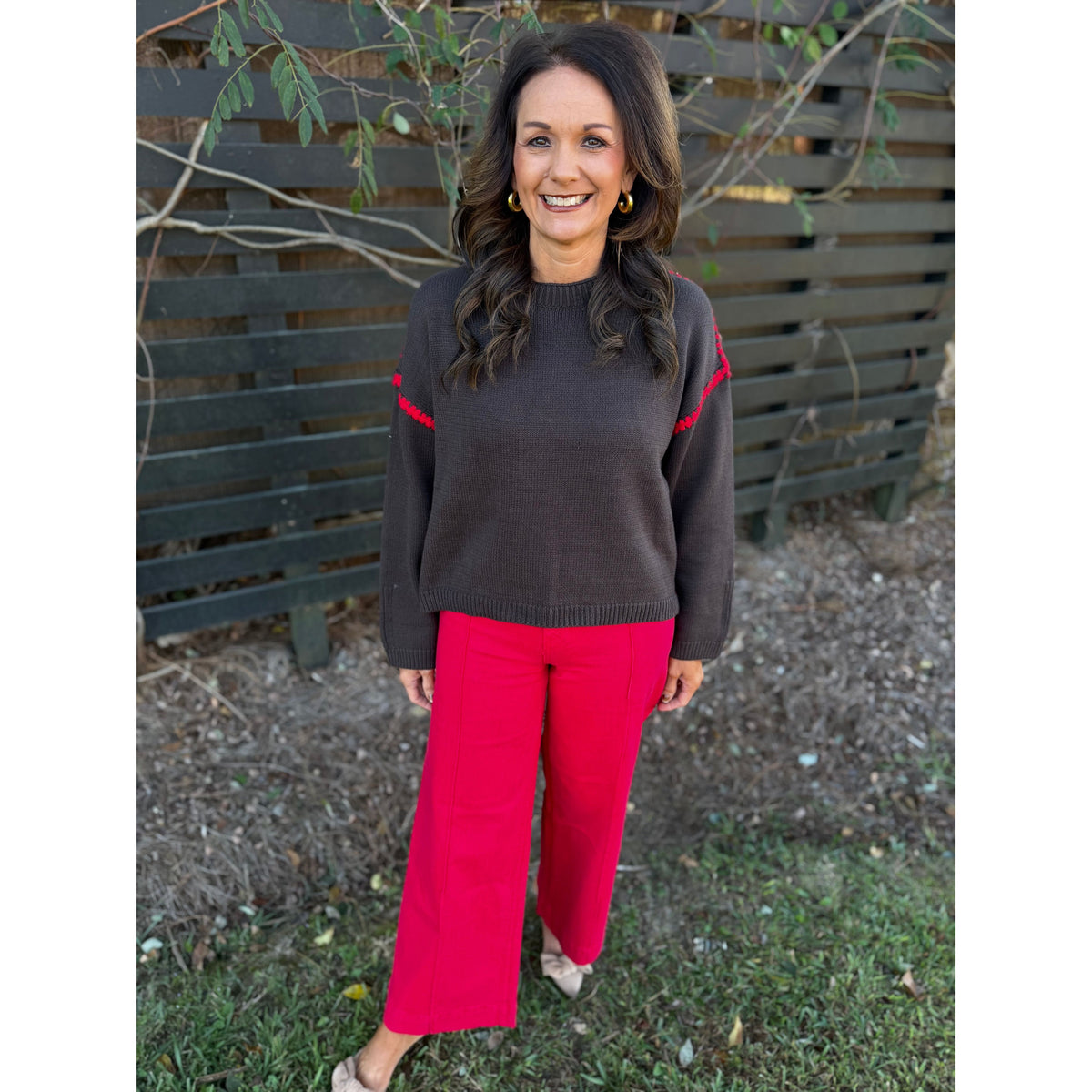 Annie Ks Boutique chocolate brown sweater with red blanket-stitch detail worn by a smiling woman in red trousers.