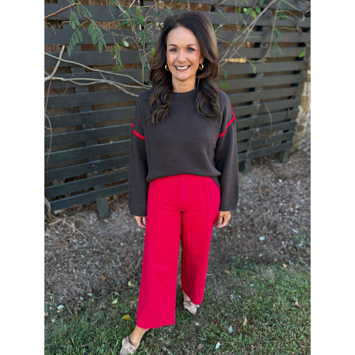 Annie Ks Boutique chocolate brown sweater with red blanket-stitch detail worn by a smiling woman with red trousers.