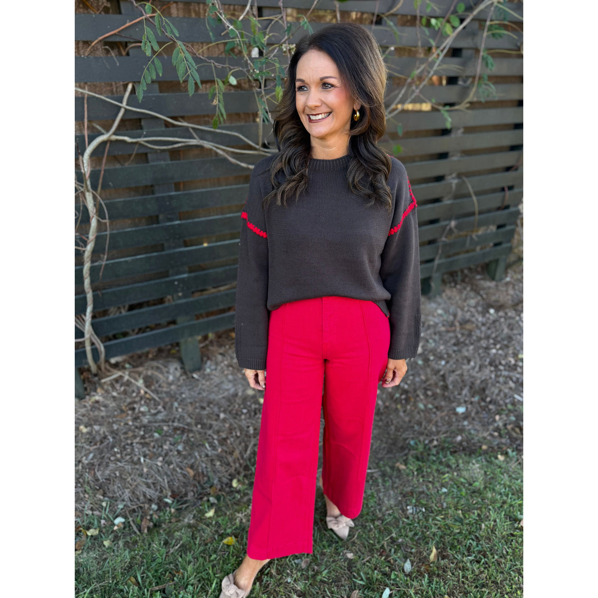Annie Ks Boutique chocolate sweater with red blanket-stitch detail worn by a smiling woman with red trousers