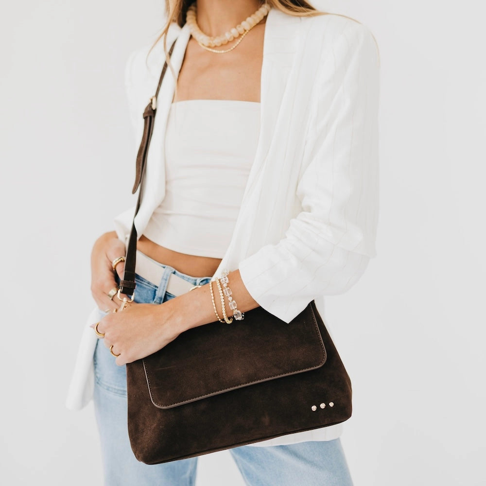 Annie Ks Boutique Miranda chocolate faux suede crossbody bag with adjustable strap worn by a woman in a white blazer