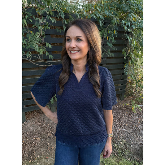 Annie Ks Boutique navy metallic textured top with ruffle collar, flutter sleeves, and smocked hem worn by a woman