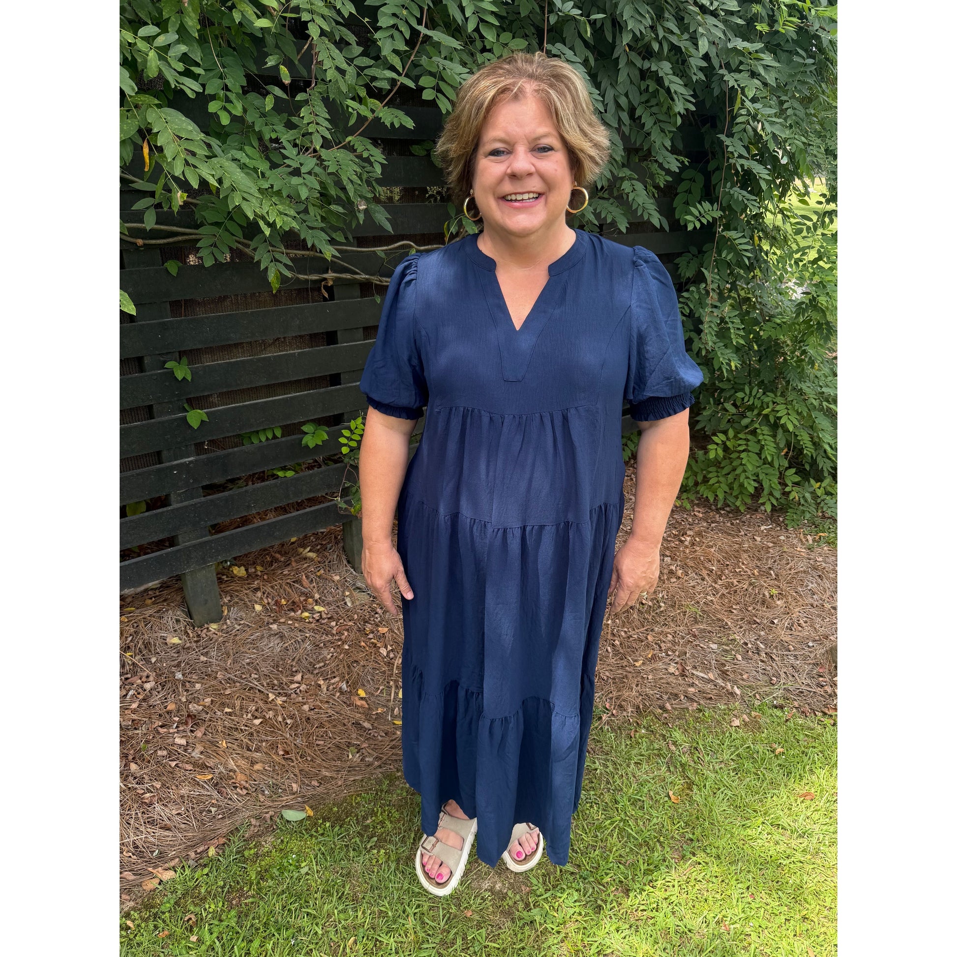 Annie Ks Boutique navy V-neck tiered midi dress with 3/4 puff sleeves worn by a smiling woman in a garden setting.