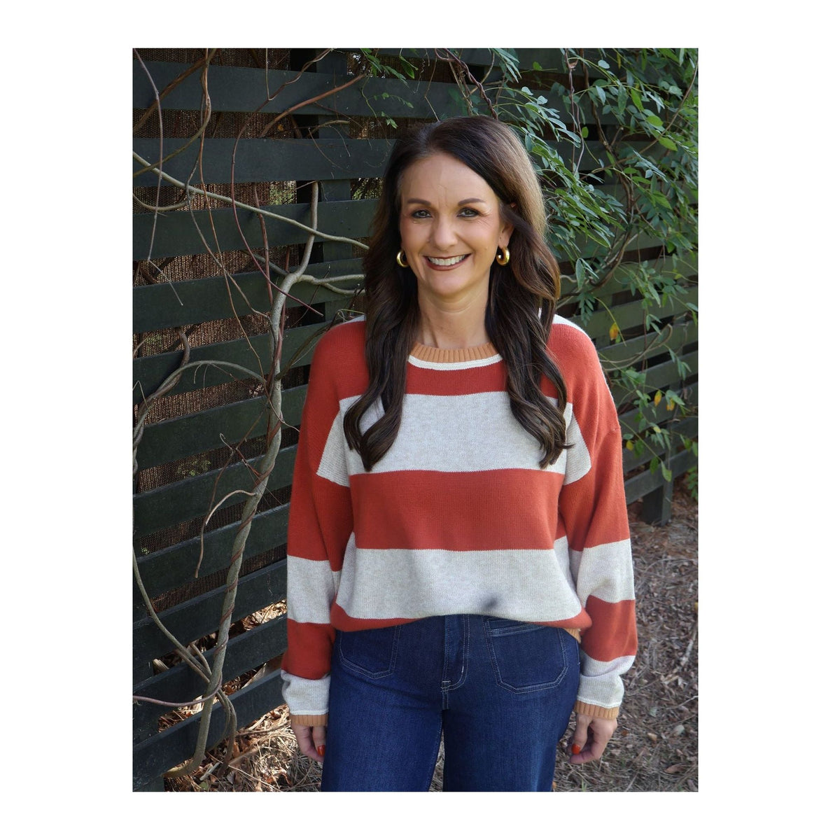 Annie Ks Boutique model wearing an oatmeal and brick red wide-striped knit sweater with a crew neck and denim jeans.