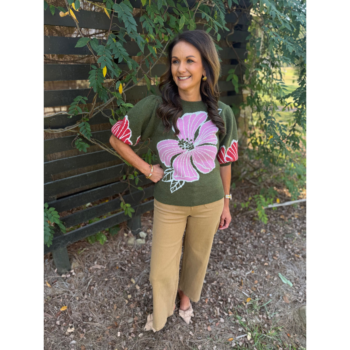 Annie Ks Boutique olive sweater with pink floral print and puff sleeves worn with tan trousers by a garden fence