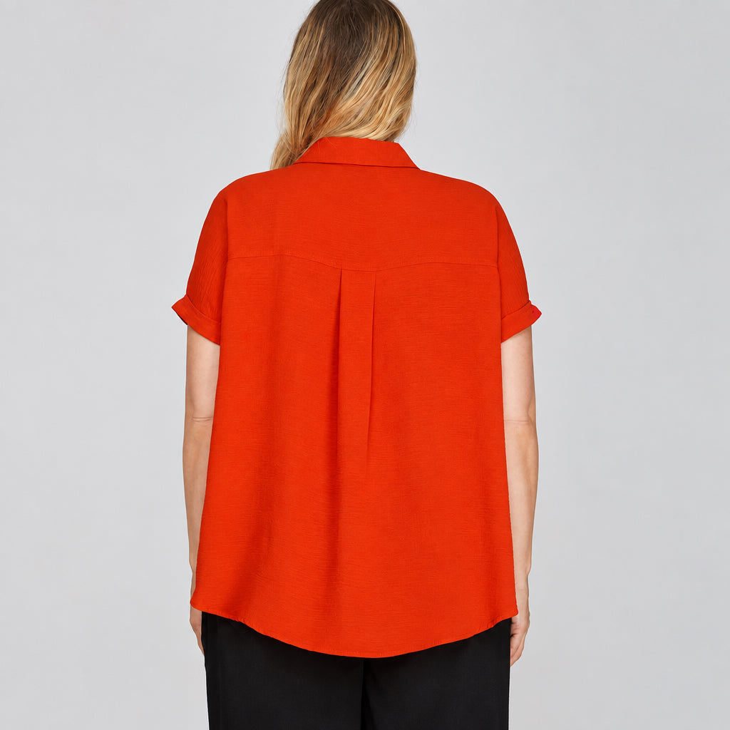 A woman viewed from the back wearing a bright orange short-sleeve collared blouse and black pants against a light gray studio background.