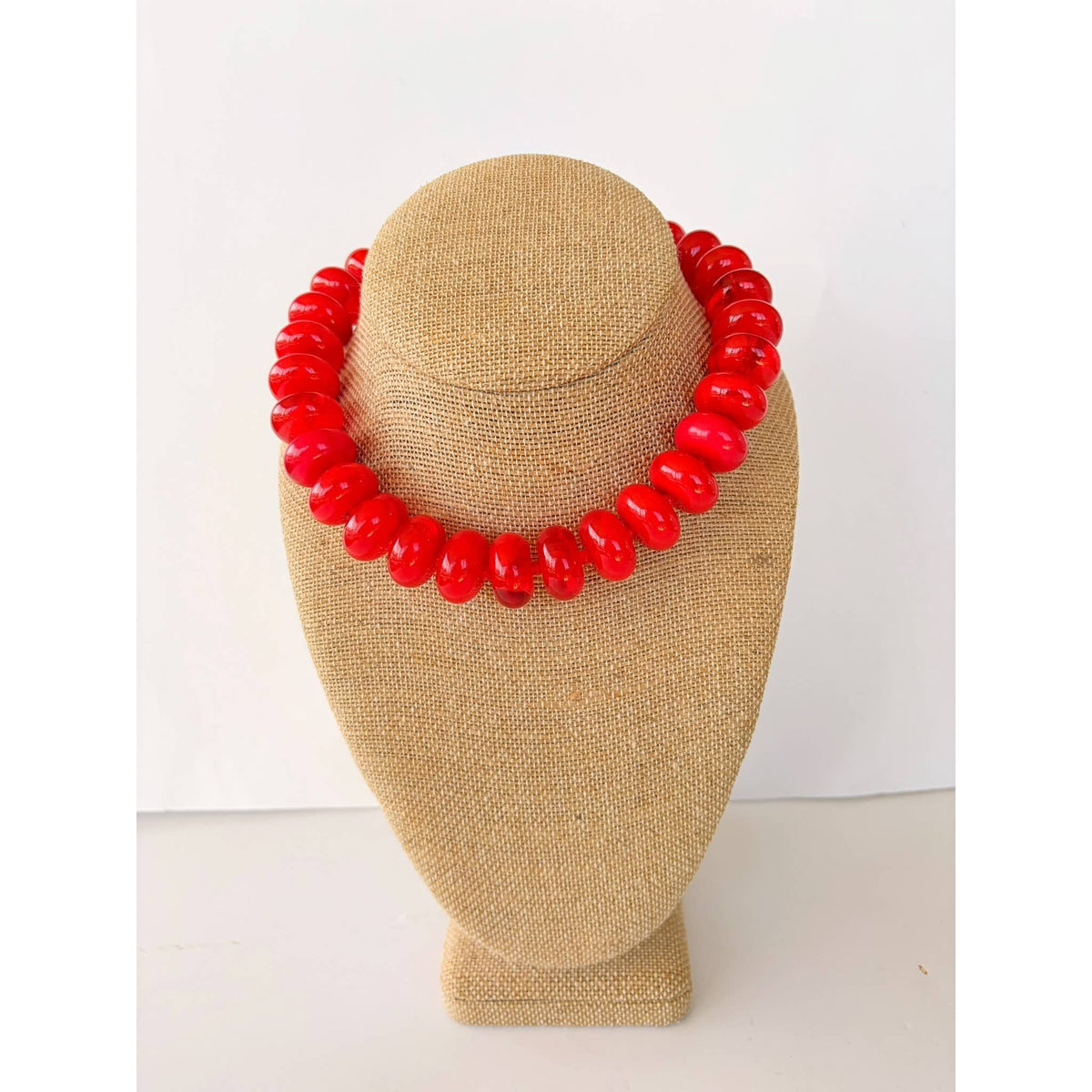 Annie Ks Boutique bright red statement necklace featuring oversized glossy rondelle beads on a beige display bust.