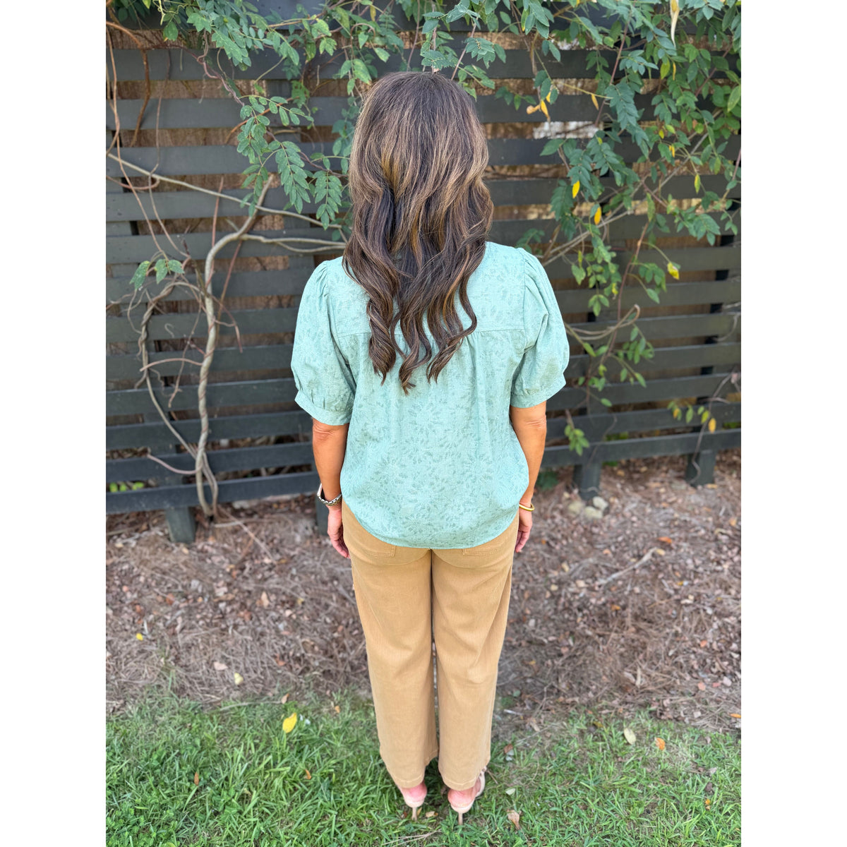 Back view of a woman wearing an Annie Ks Boutique sage puff sleeve textured blouse paired with tan trousers