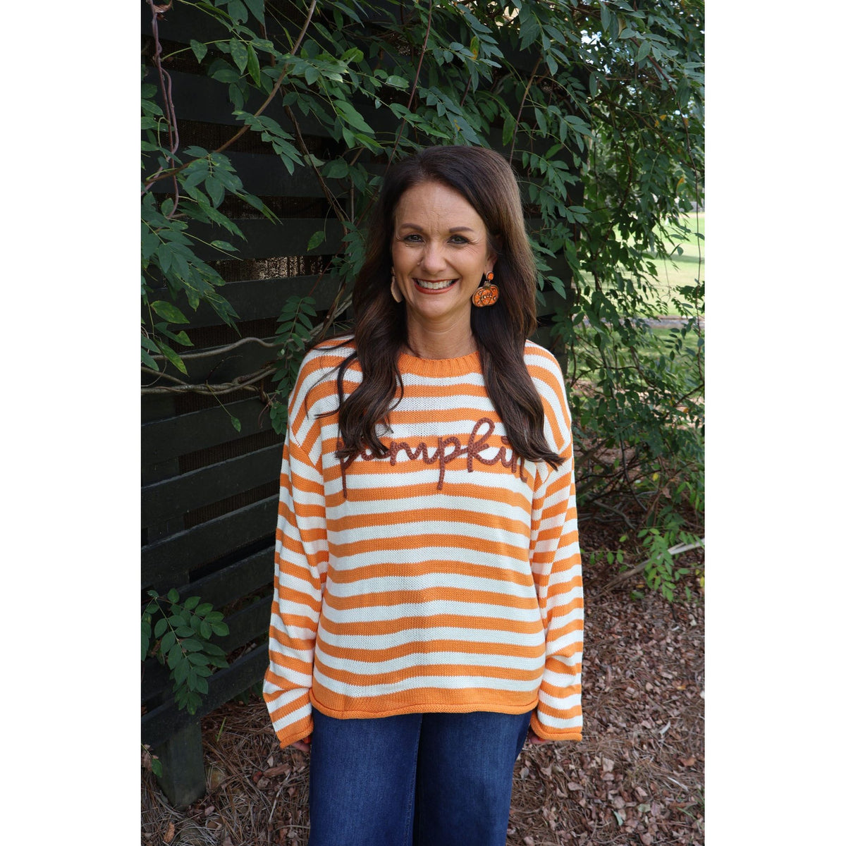Annie Ks Boutique model wearing an orange and ivory striped knit sweater with pumpkin script applique outdoors