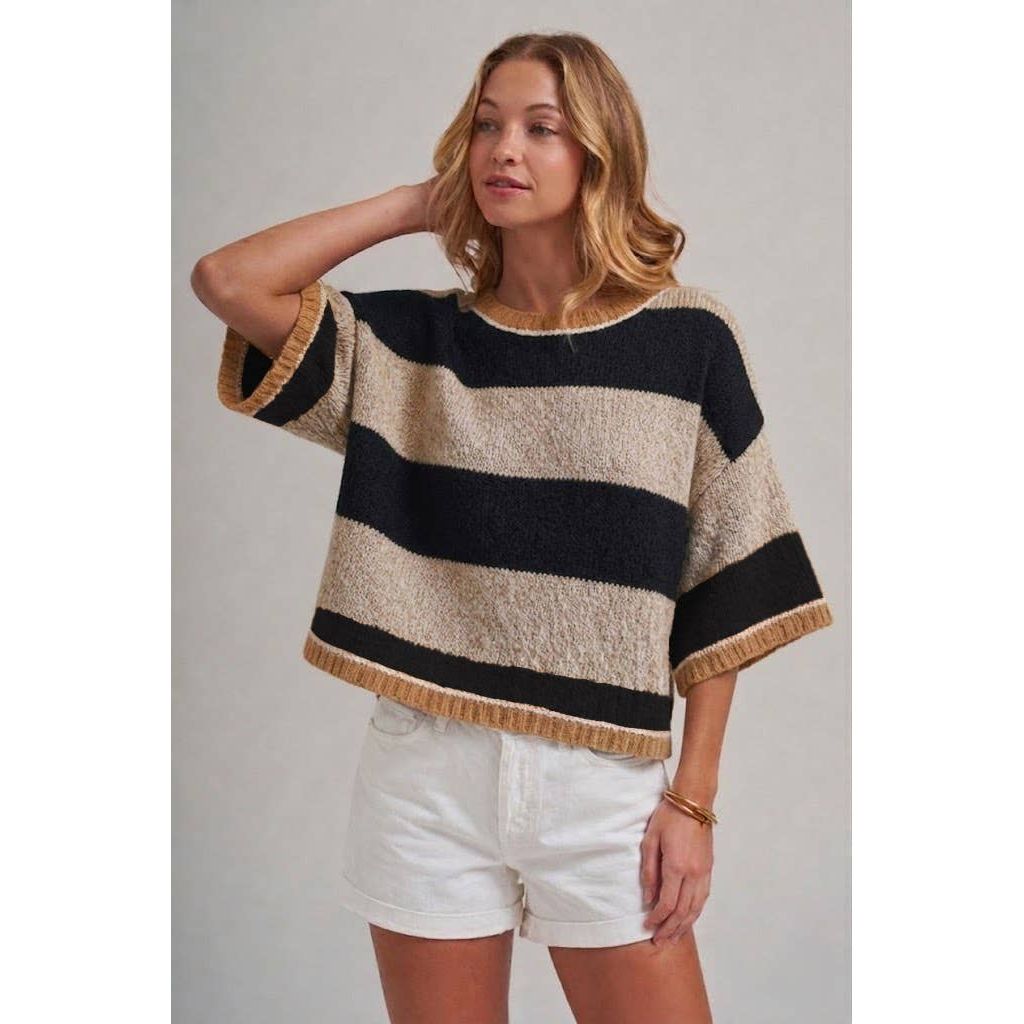A woman wearing a loose, short-sleeve knit top with wide horizontal black and beige stripes and fringed trim, paired with white shorts, against a plain light gray studio background.